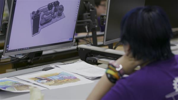 A student in the foreground looks at paper and a computer screen